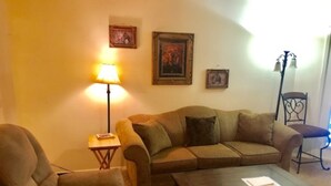 Een woonkamer met een bank, fauteuil, salontafel, lamp en lijstjes met foto’s aan de muur.