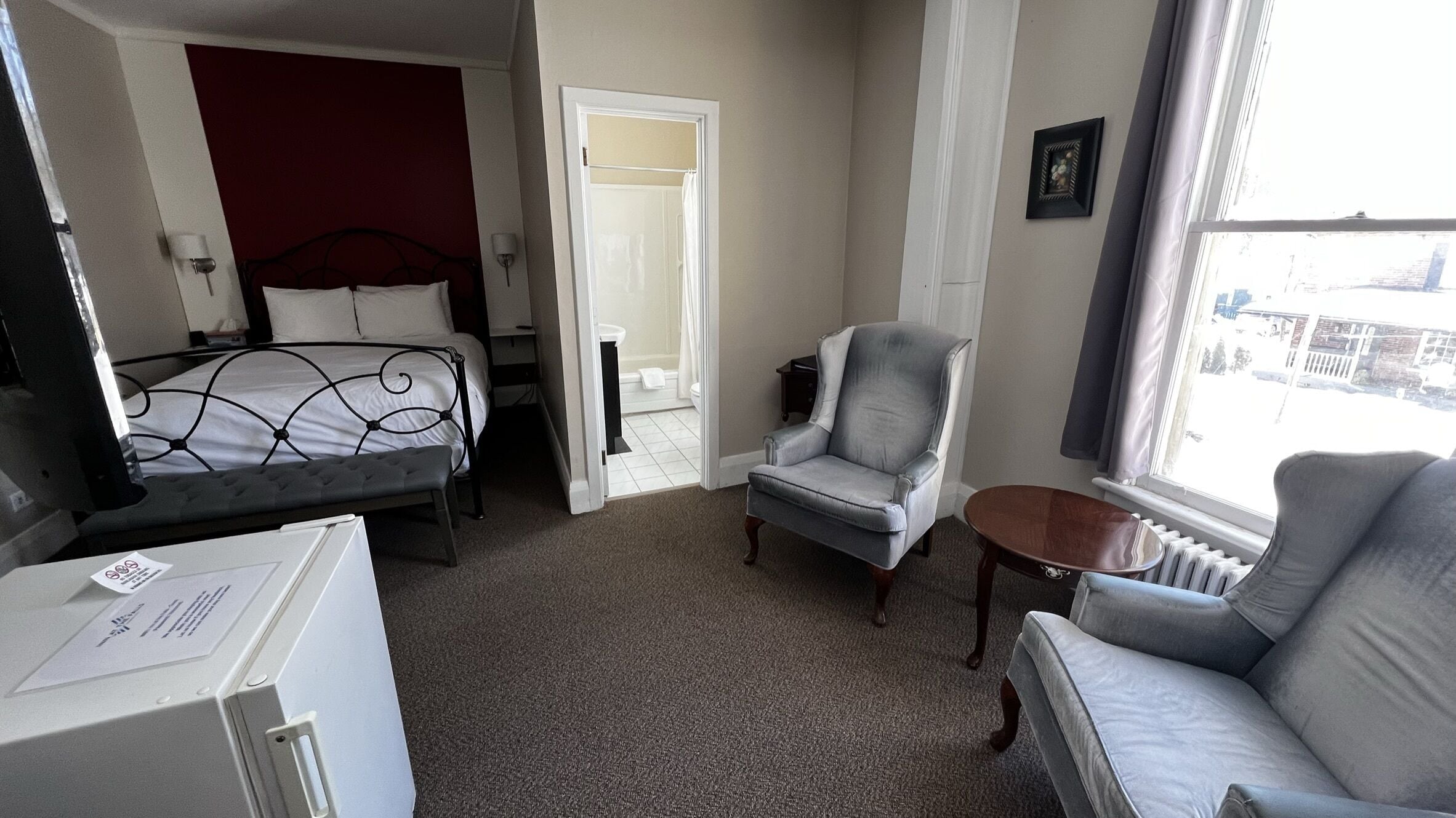 A hotel room with a bed, a grey armchair, a small table, and a window with a view of buildings.