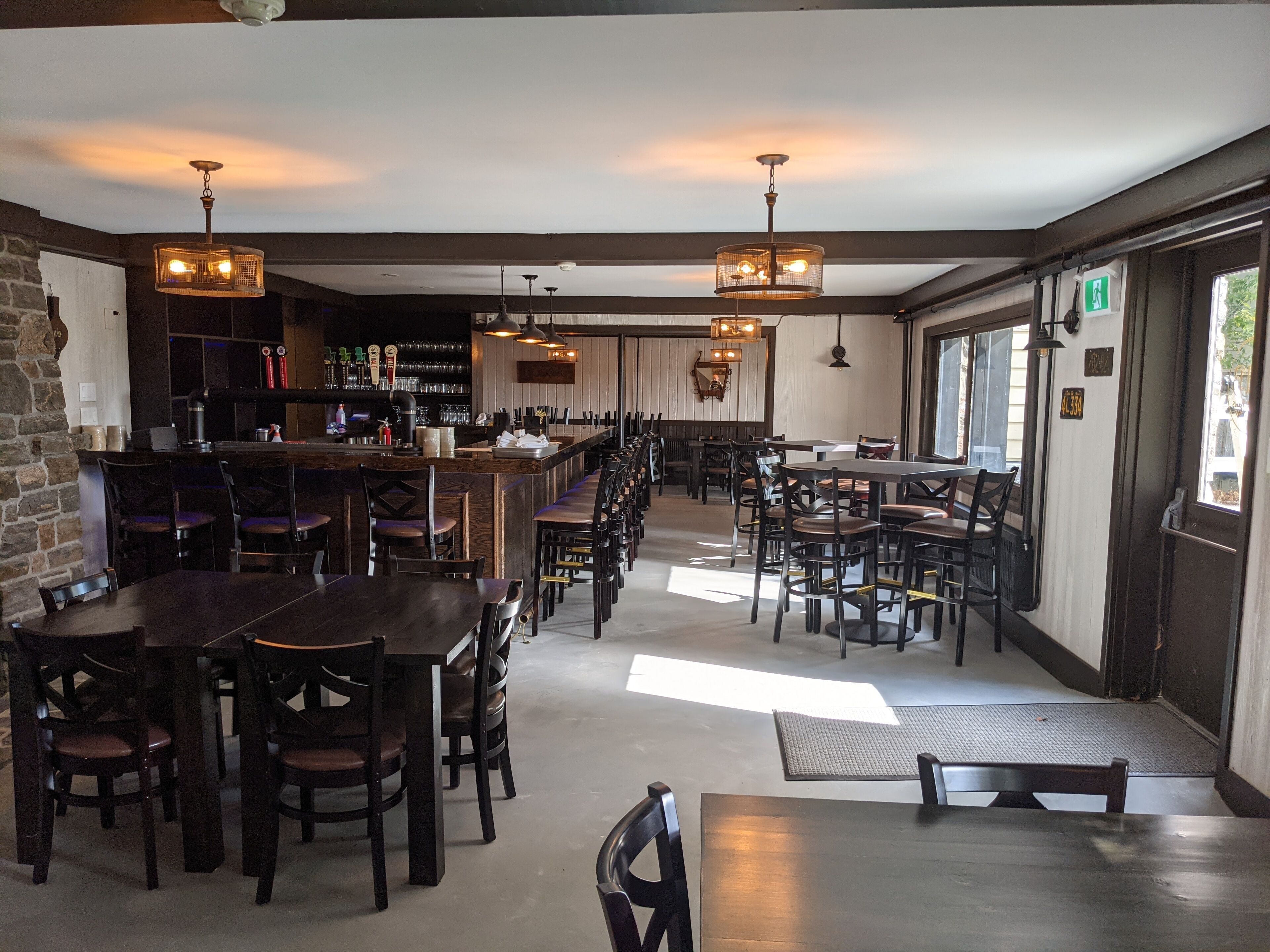 A restaurant with a long counter, multiple tables, and chairs. There are hanging lights and a bar area with bottles and glasses.