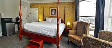 A four-poster bed with a red frame, a small wooden step stool, a sofa, a desk with a microwave, and a television.