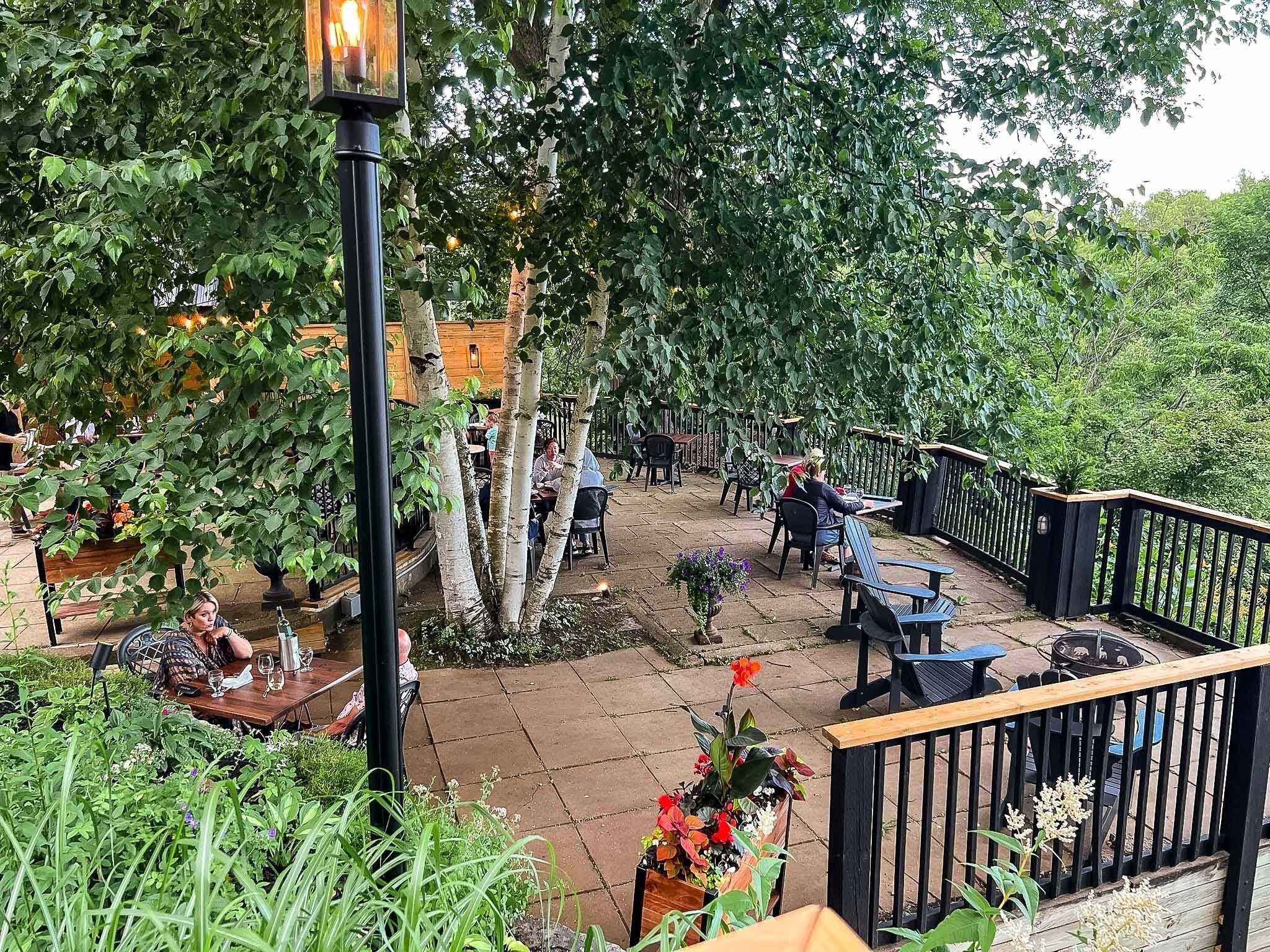 Un espace extérieur aménagé en terrasse, avec des tables et des chaises, entouré d’une végétation luxuriante et d’arbres.