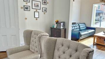 Una sala de estar con un sofá color beige, un sofá azul y una mesa de centro de madera. Se observan cuadros enmarcados en la pared y una ventana con vistas al exterior.