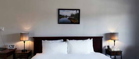 A neatly made bed with white linens, two bedside tables with lamps, and a framed landscape painting on the wall.