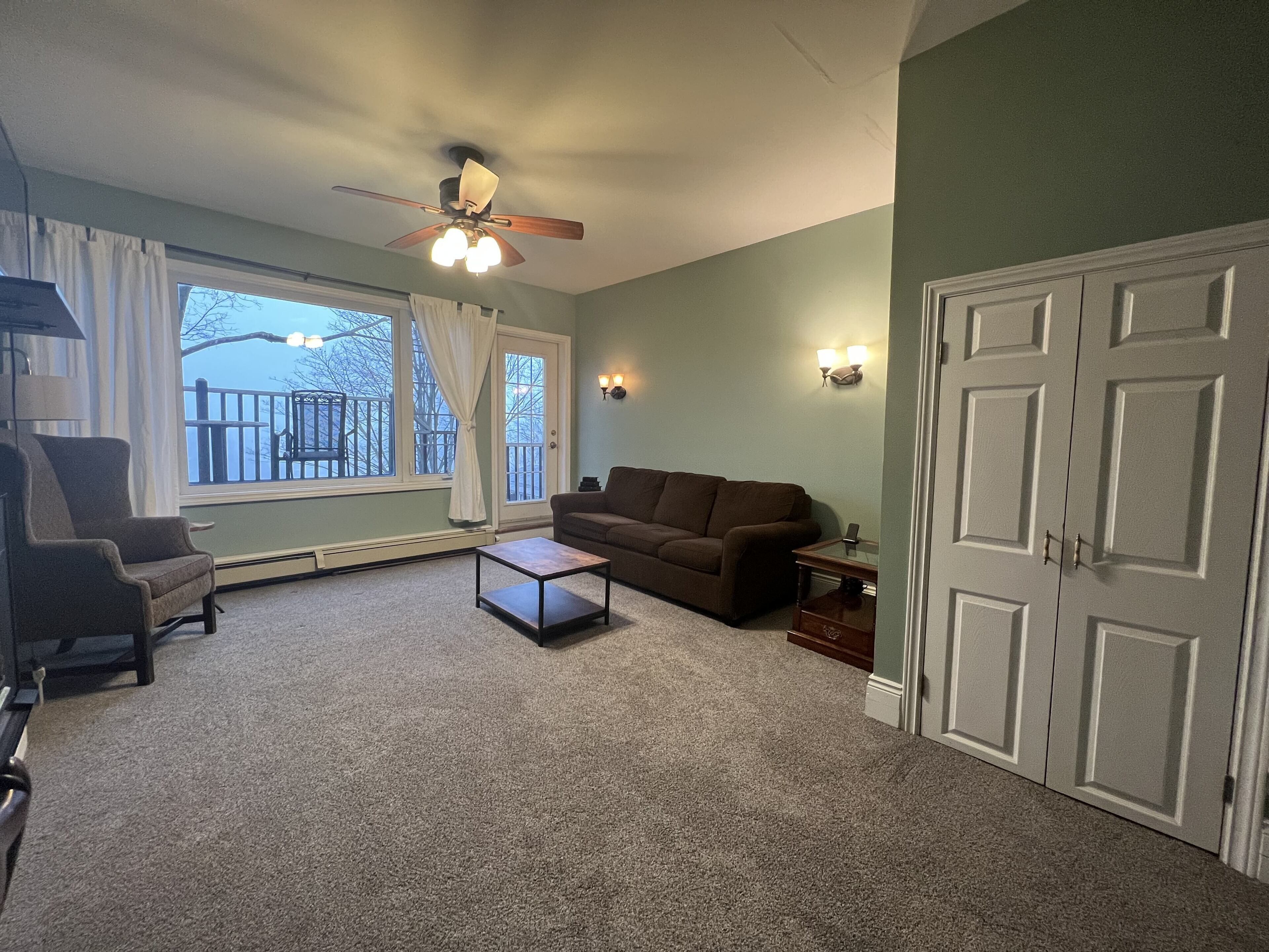 A living room with a brown sofa, a small coffee table, a chair, a ceiling fan, and a view of a balcony with a railing.