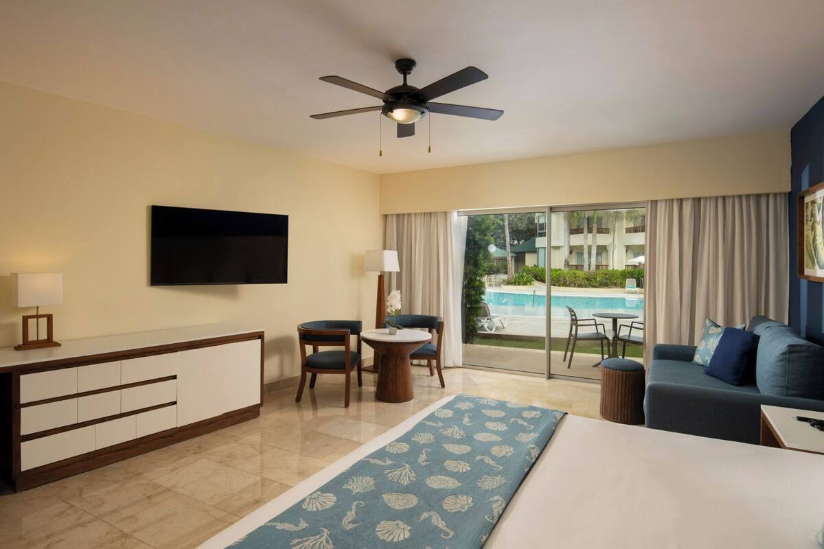 Ein Hotelzimmer mit Bett, Schreibtisch, Sessel, Fernseher, Deckenventilator und Blick auf einen Pool.
