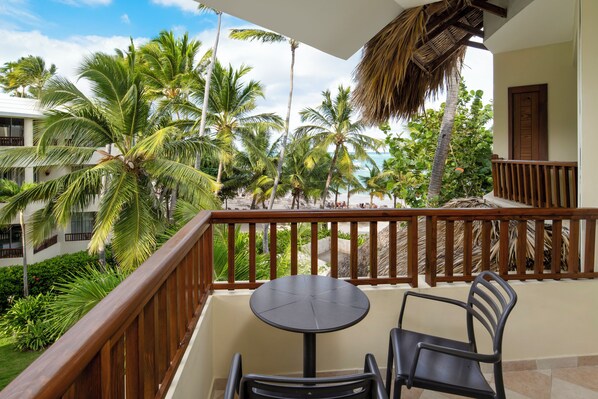 A balcony with a table and chairs, overlooking a beach with palm trees and a thatched roof structure.