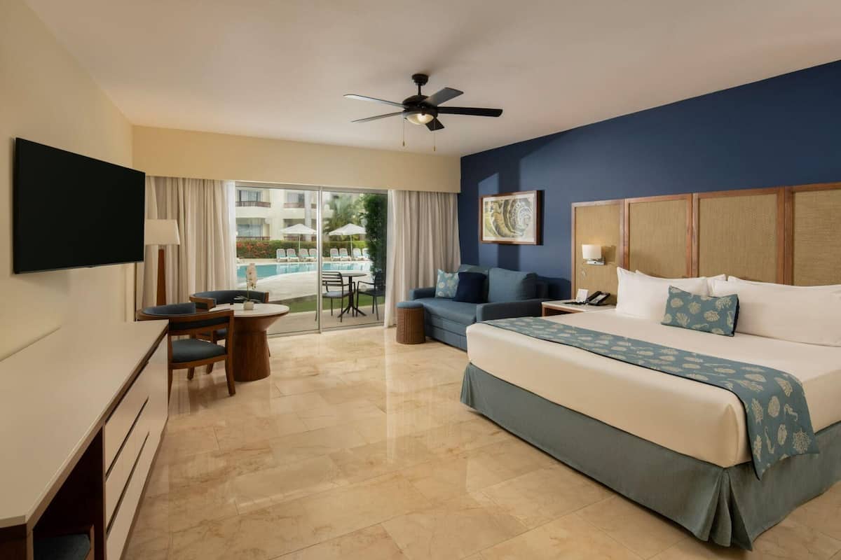 Ein Hotelzimmer mit Bett, Schreibtisch, Stuhl, Fernseher, Deckenventilator und Blick auf einen Pool.