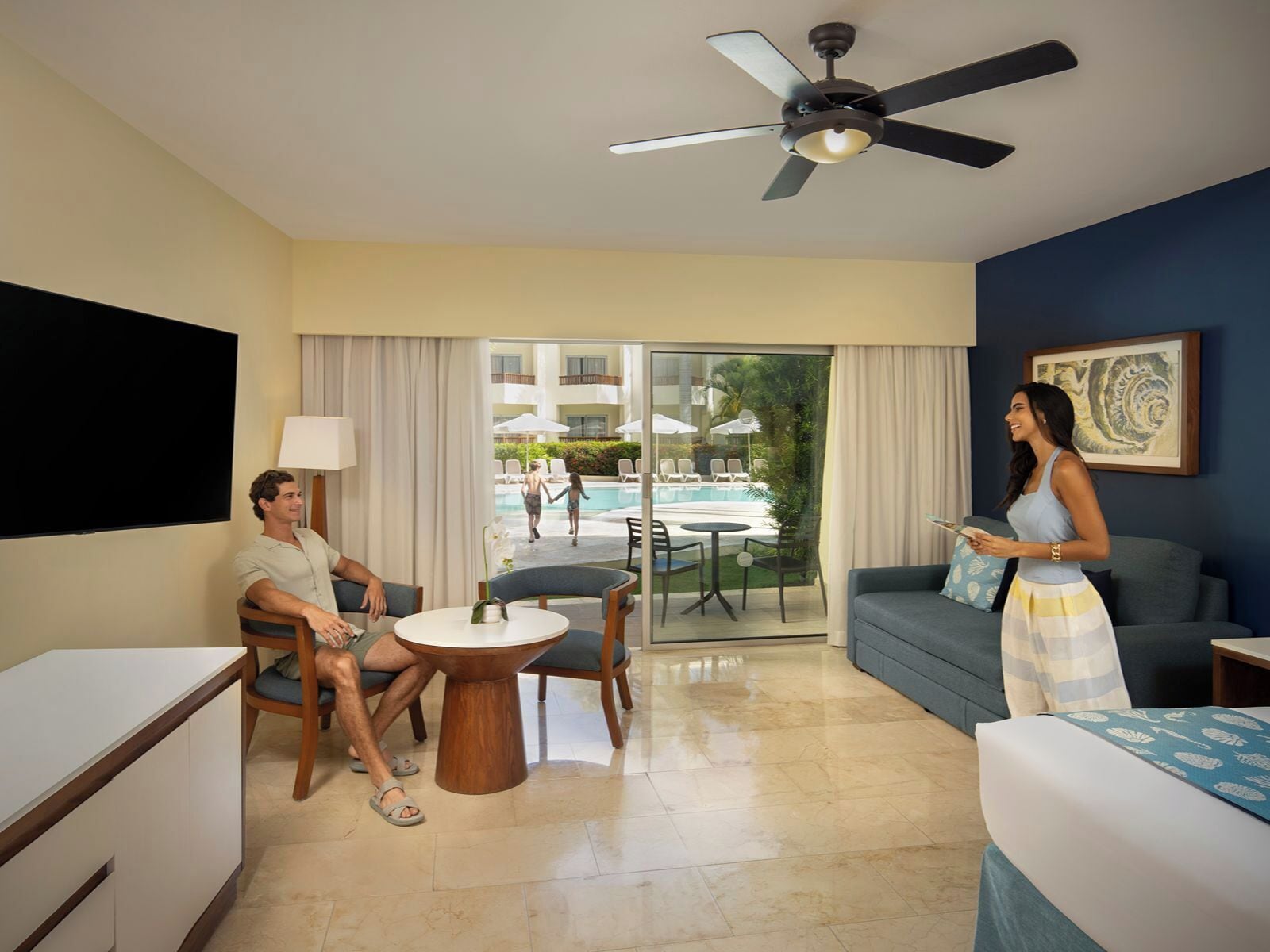 A hotel room with a balcony, a ceiling fan, a TV, a dining table, and a sofa.