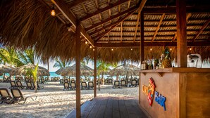 A beach bar with wooden deck and thatched roof, surrounded by beach chairs and umbrellas. - Impressive Premium Punta Cana (Punta Cana)