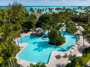 Une piscine de complexe hôtelier aménagée dans un décor tropical, entourée de palmiers et offrant une vue sur l’océan.