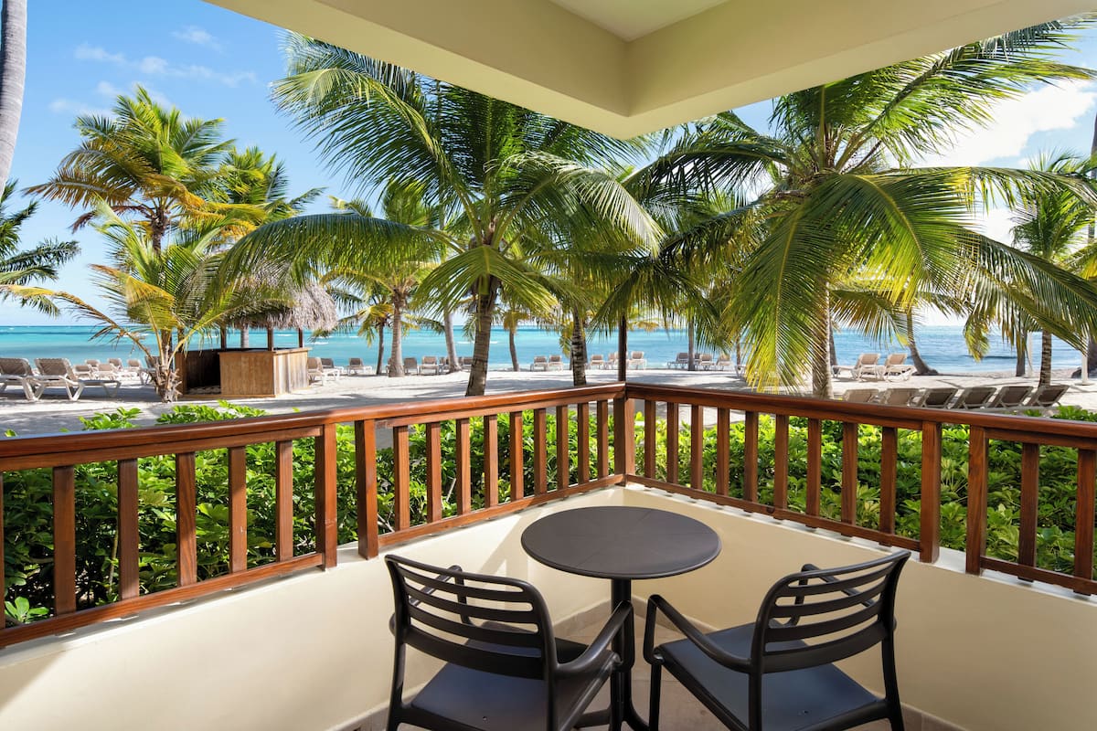 Ein Balkon mit zwei Stühlen und einem Tisch, der Blick auf einen Strand mit Palmen und Liegestühlen bietet.