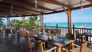 An outdoor dining area with wooden tables and wicker chairs, overlooking a beach with palm trees and the ocean. - Impressive Premium Punta Cana (Punta Cana)