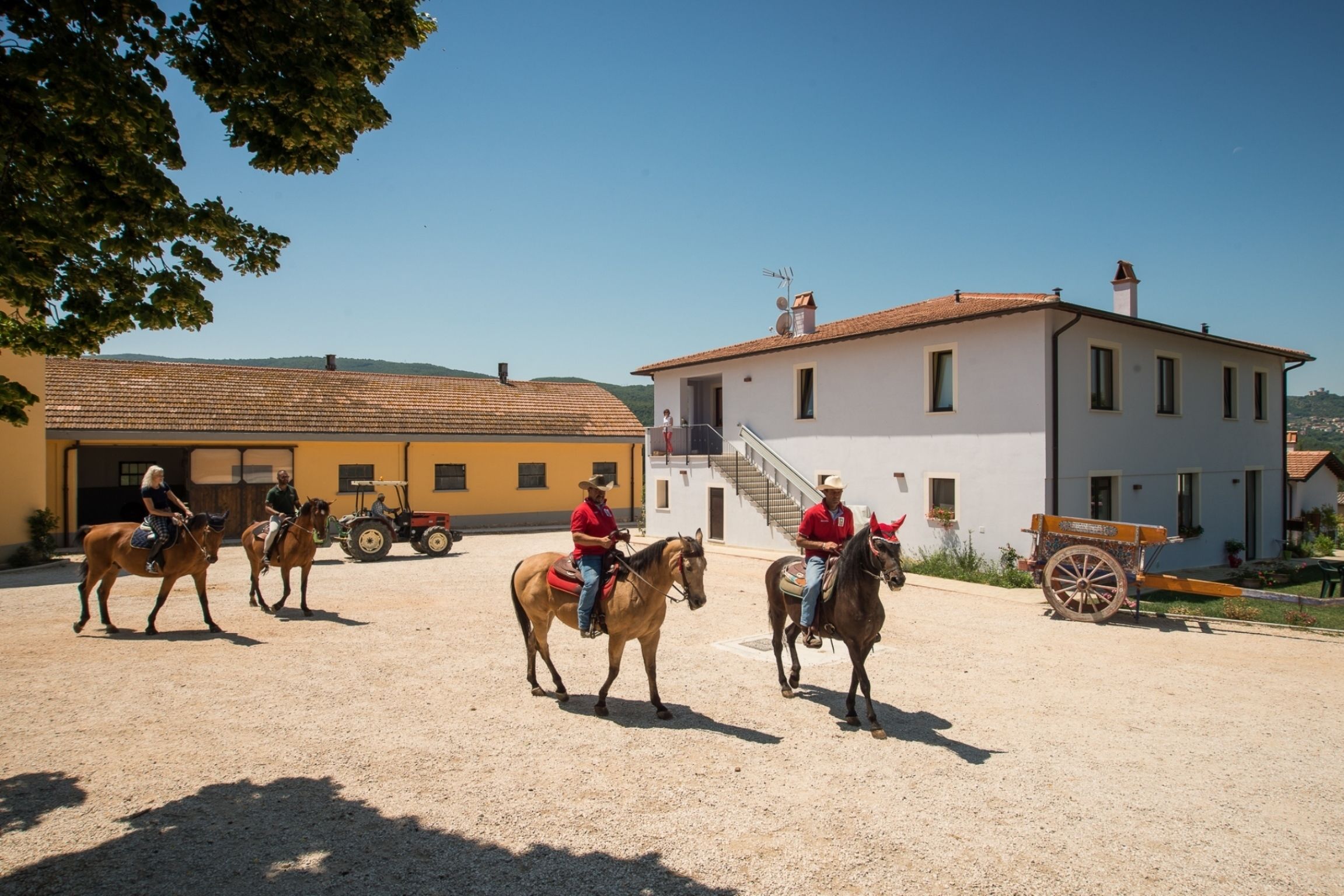 Een stal met paarden en ruiters, een gebouw met een rood dak, en een heldere lucht.