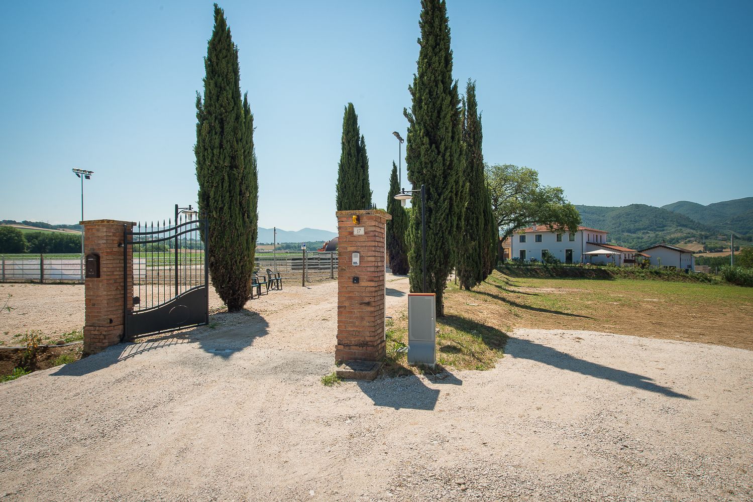 Un ingresso recintato con alti cipressi, un vialetto sterrato e un edificio con tetto rosso sullo sfondo.