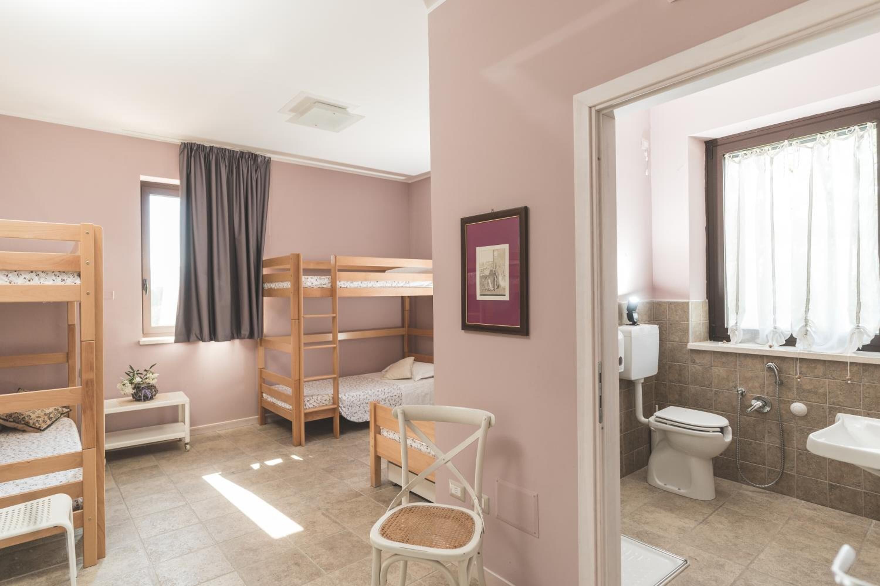 A room with bunk beds, a small table with flowers, a chair, and a bathroom with a toilet and sink.
