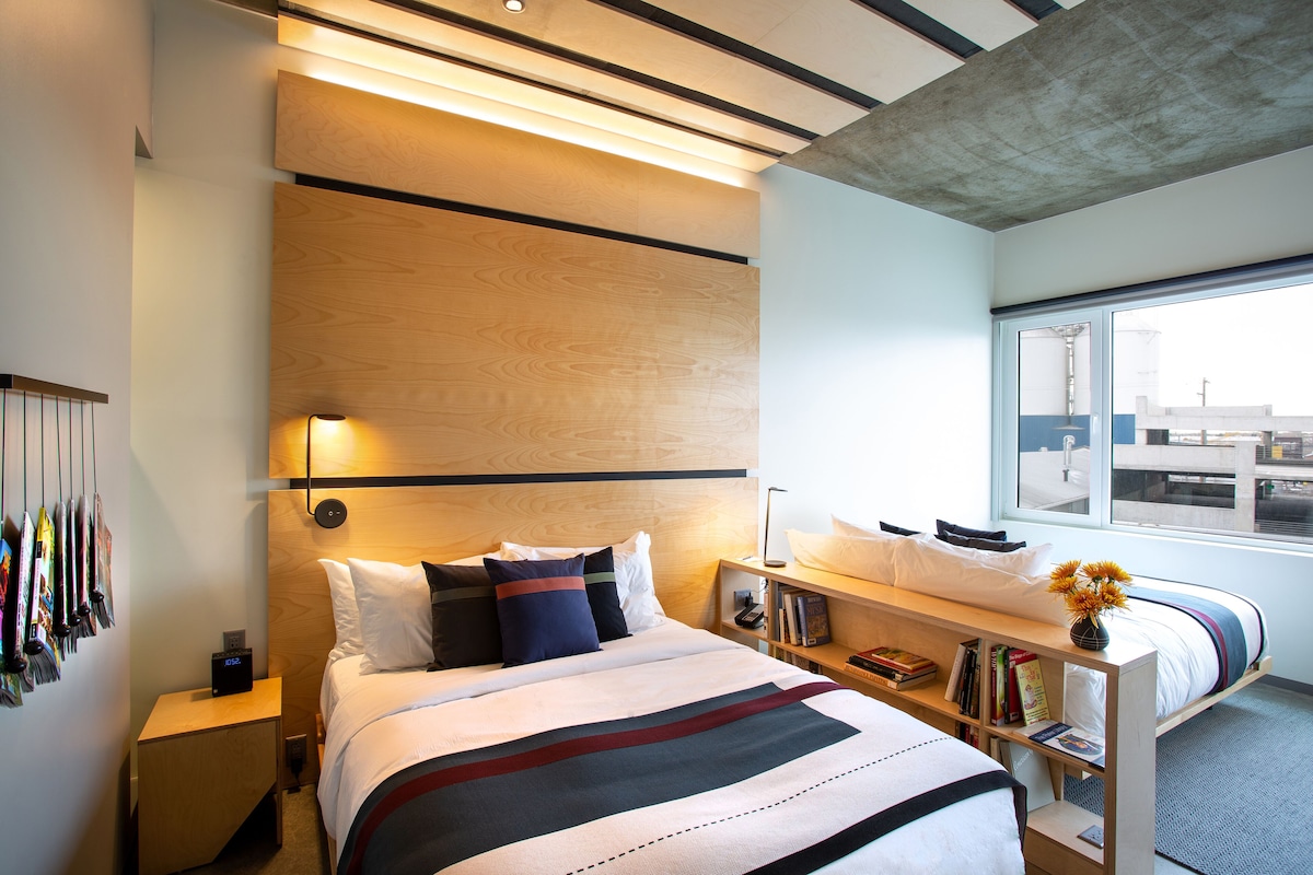 A modern hotel room with a large bed, bedside tables, a reading area, and a view of the city through the window.