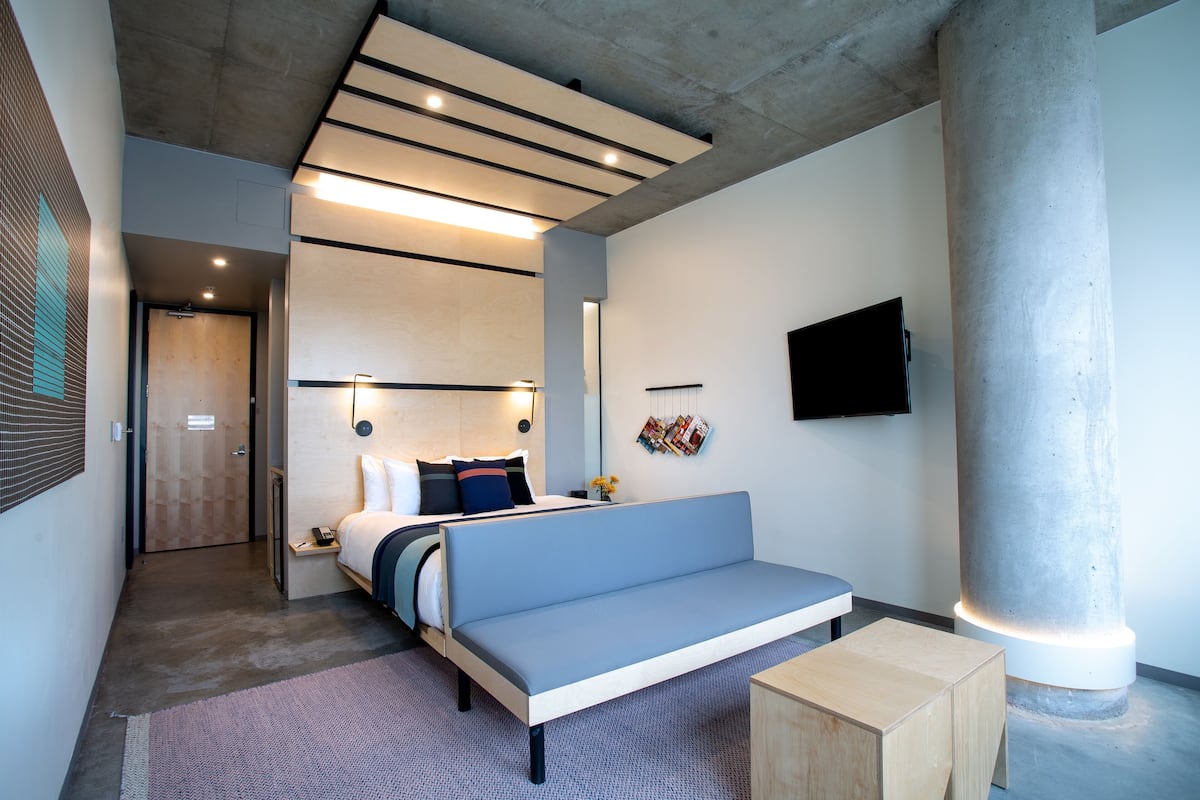 A modern hotel room with a bed, a sofa, a small wooden table, and a television mounted on the wall.