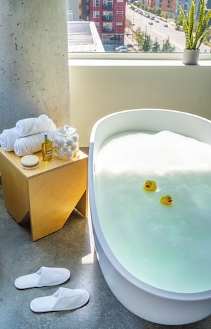 A bathroom with a bathtub, a wooden side table with towels and toiletries, and a view of the city through a window.