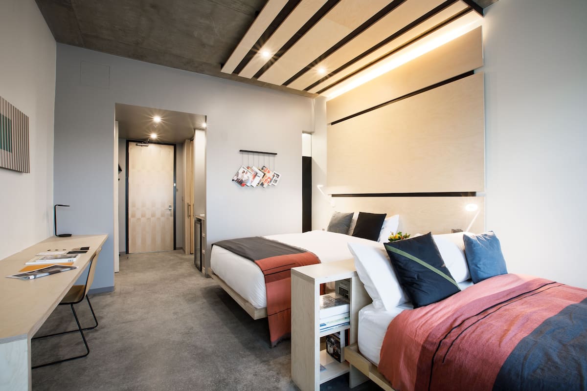 A modern bedroom with a bed, bedside table, and a desk with a chair. There is a door leading to another room.