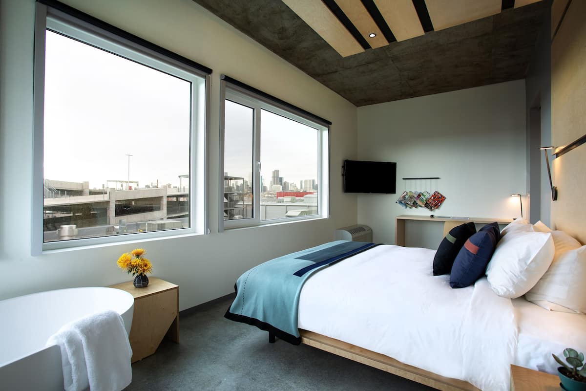 Un dormitorio moderno con una cama grande, una mesita de noche con un jarrón de flores, una ventana con vistas a la ciudad y un televisor montado.