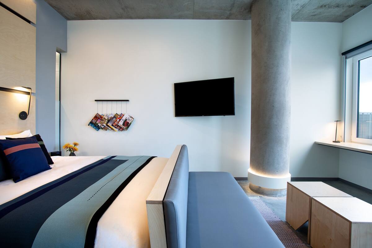 A modern hotel room with a large bed, a flat-screen TV, a wooden bench, and a window.