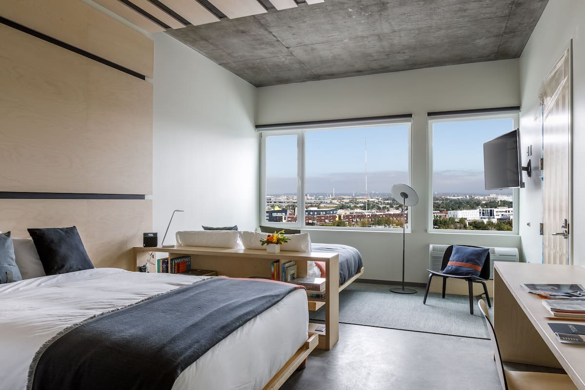 A modern hotel room with a large bed, a desk, and a chair. There are two windows offering a city view.
