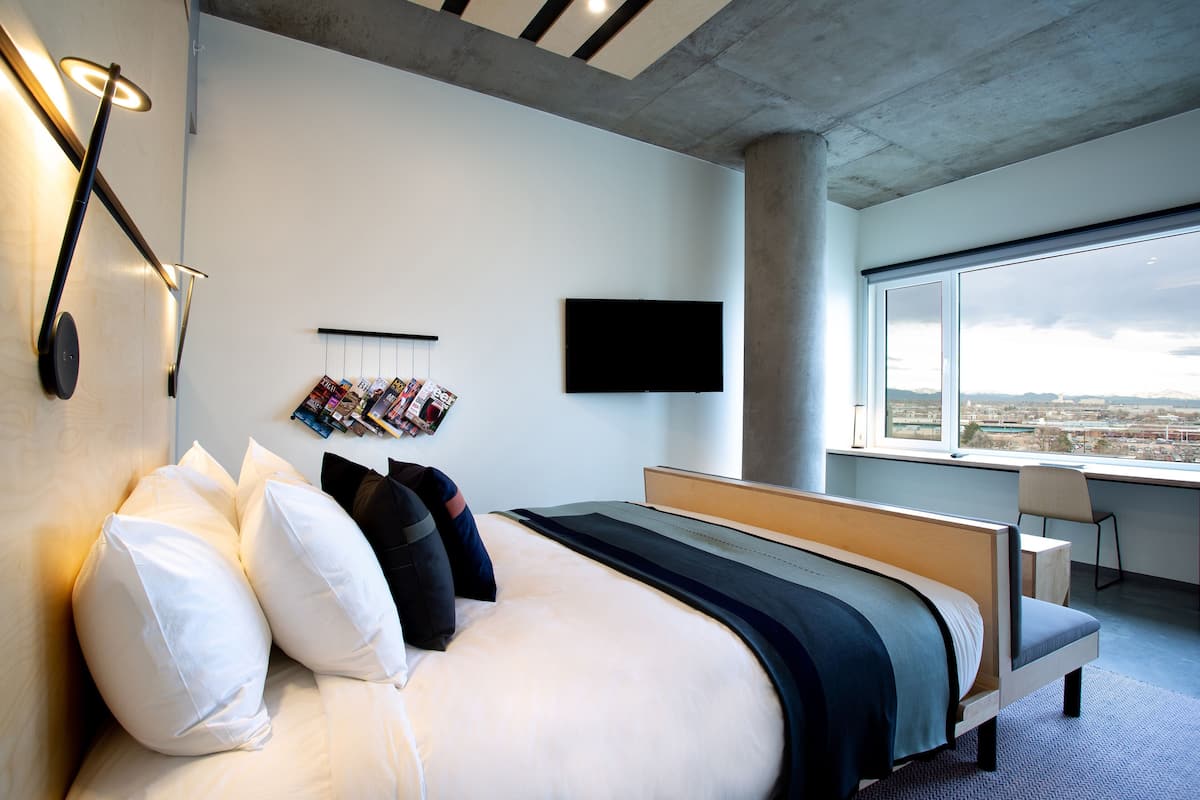 A modern hotel room with a large bed, a flat-screen TV, a window with a city view, and a desk with a chair.