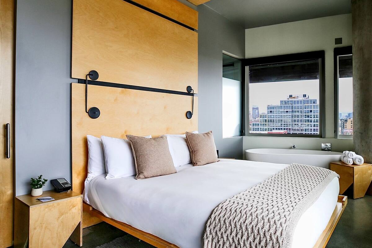 A modern hotel room with a large bed, a wooden headboard, a built-in bathtub, and a view of the cityscape through a large window.