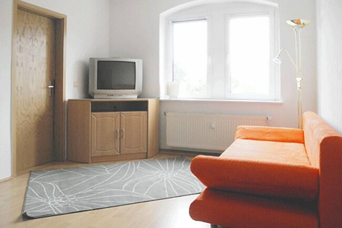 Quarto com porta de madeira, televisão sobre um armário, janela com vista e um sofá laranja.