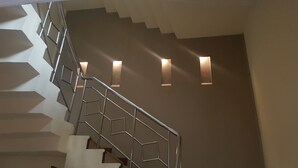 Una escalera con barandilla metálica y diseño geométrico en la pared, que conduce a un rellano con tres luces fijadas a la pared.