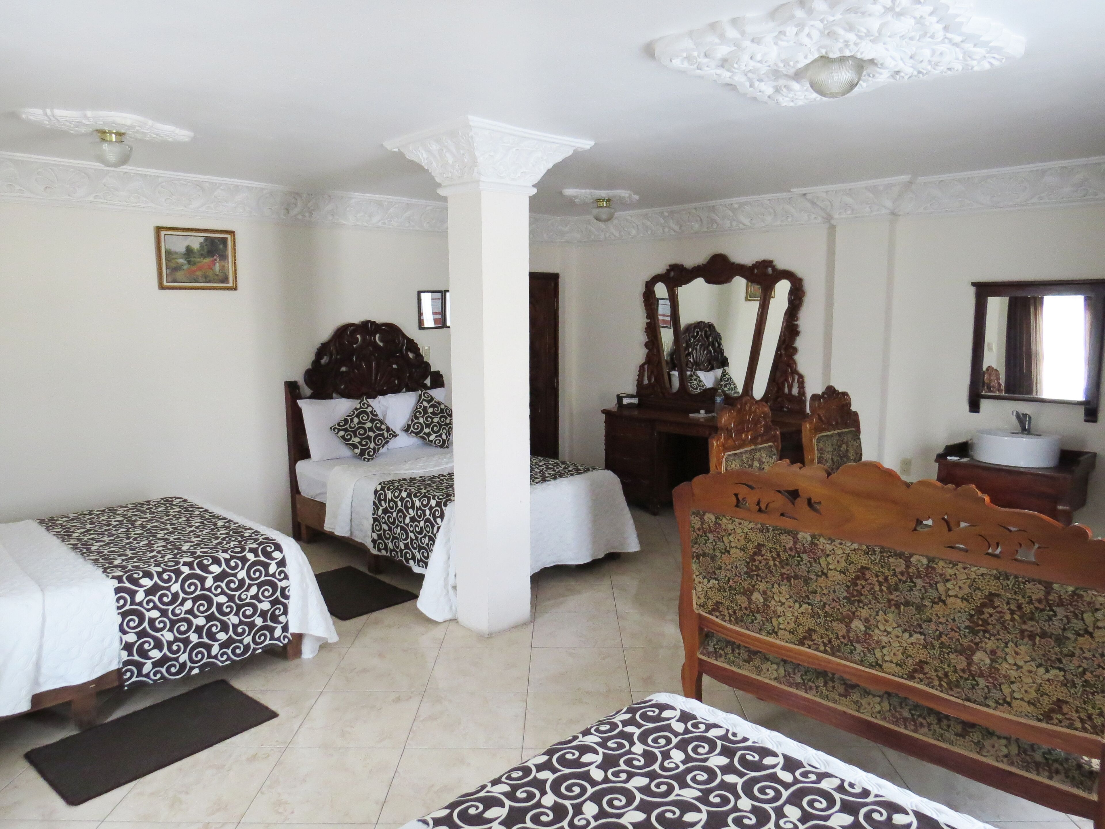 Una camera d'albergo con due letti singoli, una sedia di legno, una scrivania con uno specchio, un mobile bagno con un lavandino e una ventola a soffitto.