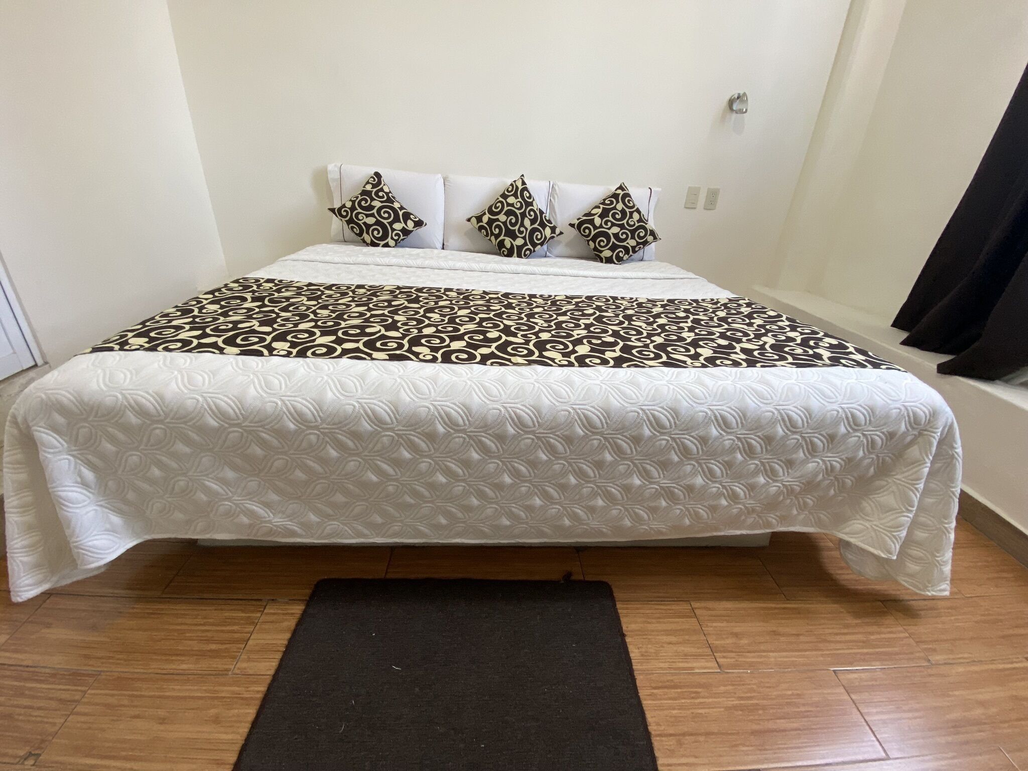 A neatly made bed with a patterned bedspread and pillows, a black rug in front, and wooden flooring.