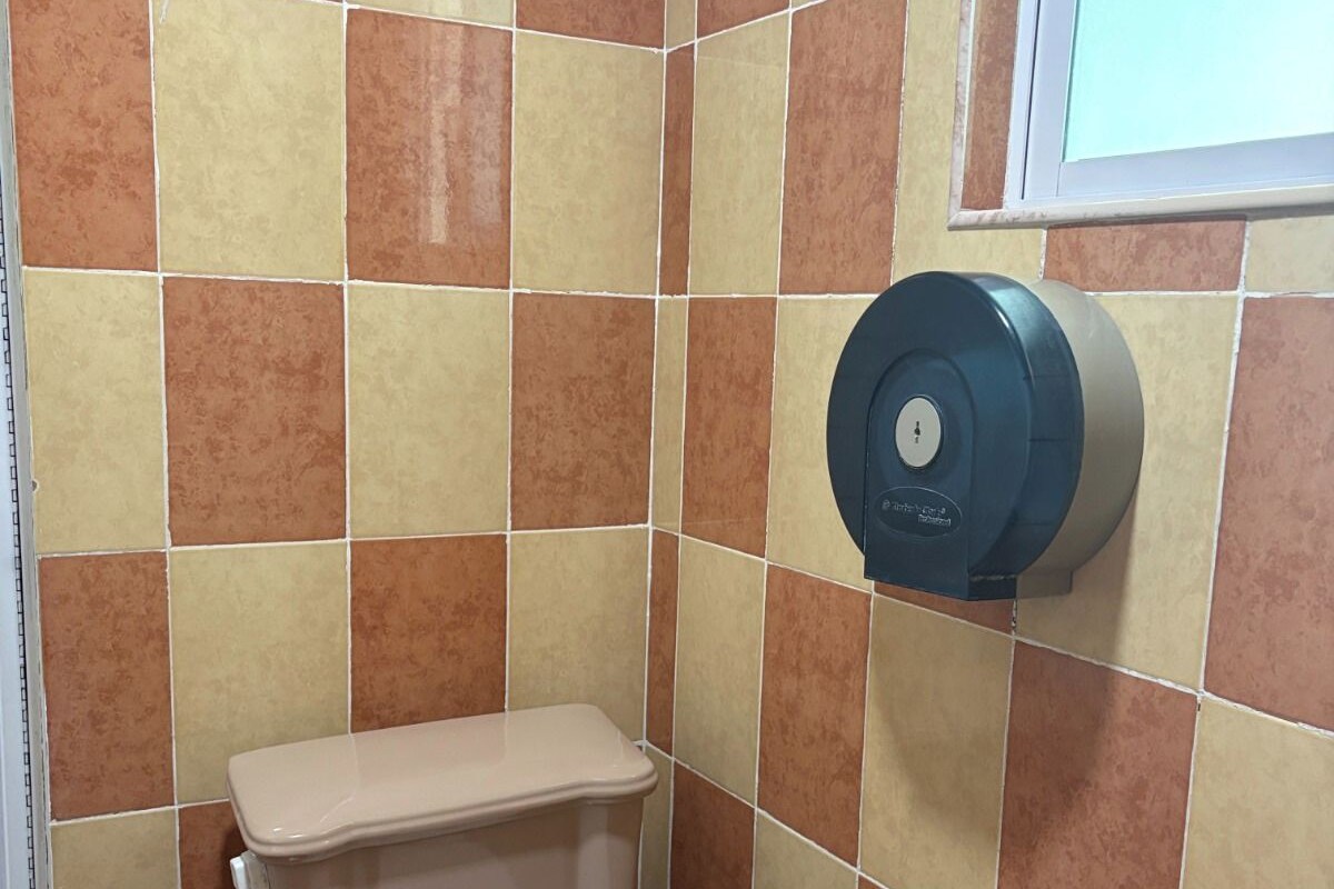 A toilet with a white seat and tank, a black trash bin, and a hand sanitizer dispenser on the wall.