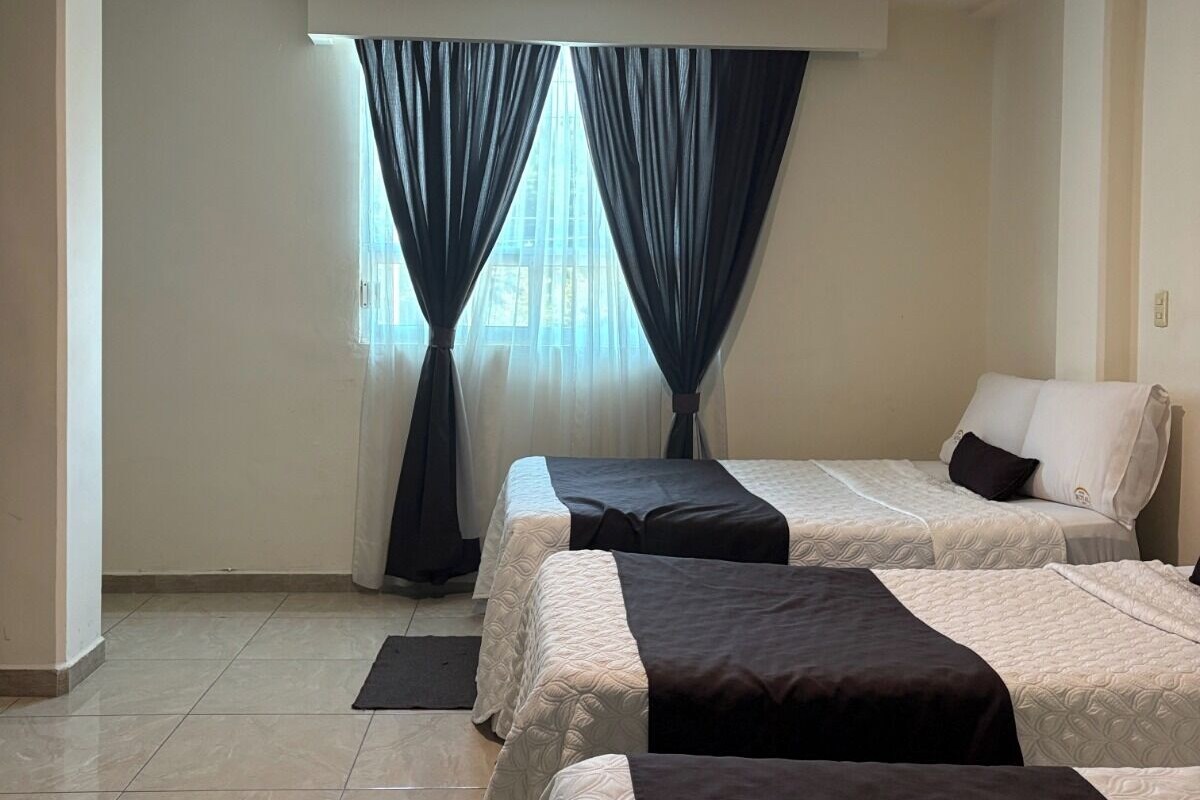 A hotel room with two beds, a ceiling light, a window with curtains, and a tiled floor.