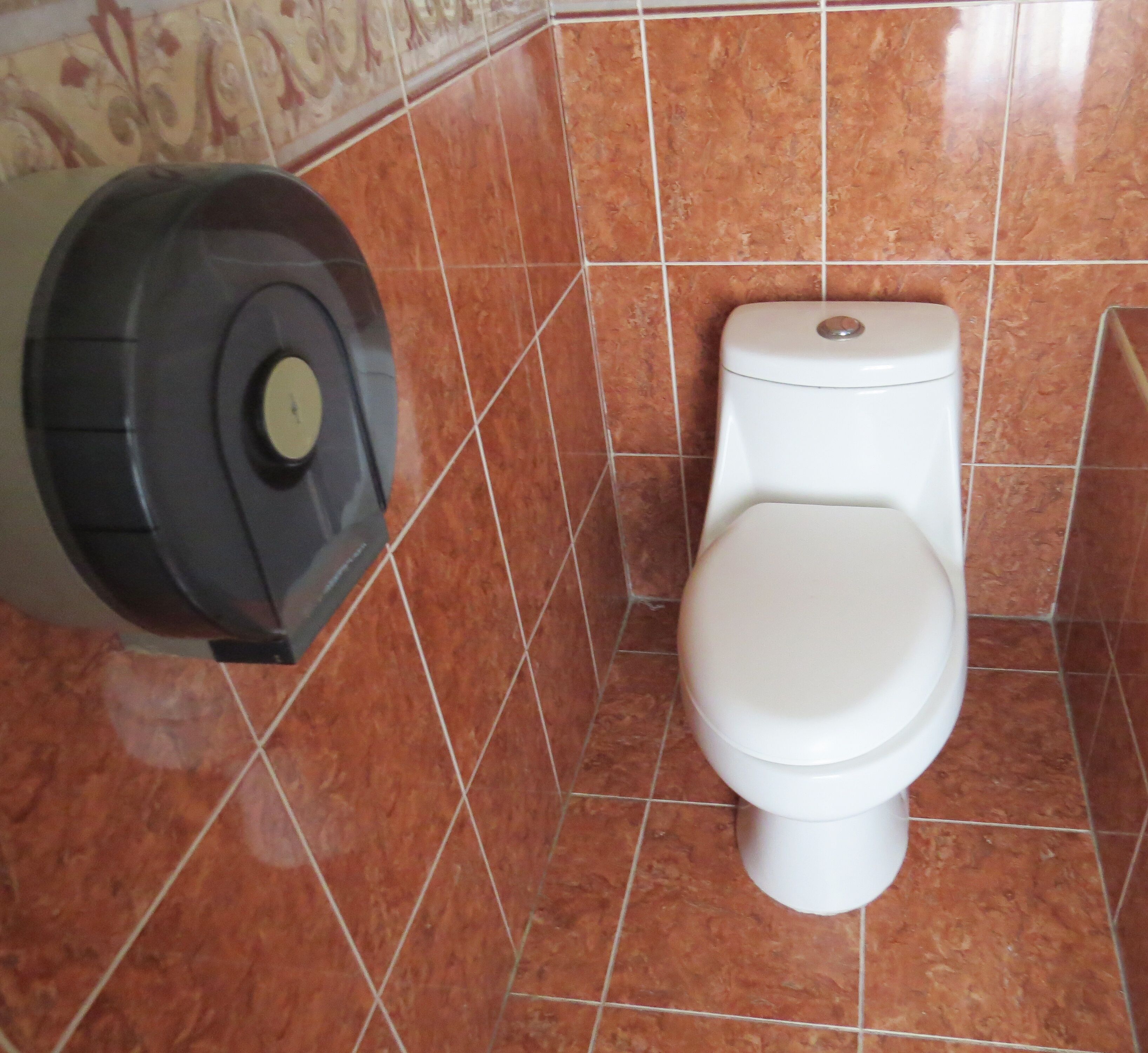 Un inodoro con bidé de pared y tapa blanca en un baño con paredes de azulejos marrones.