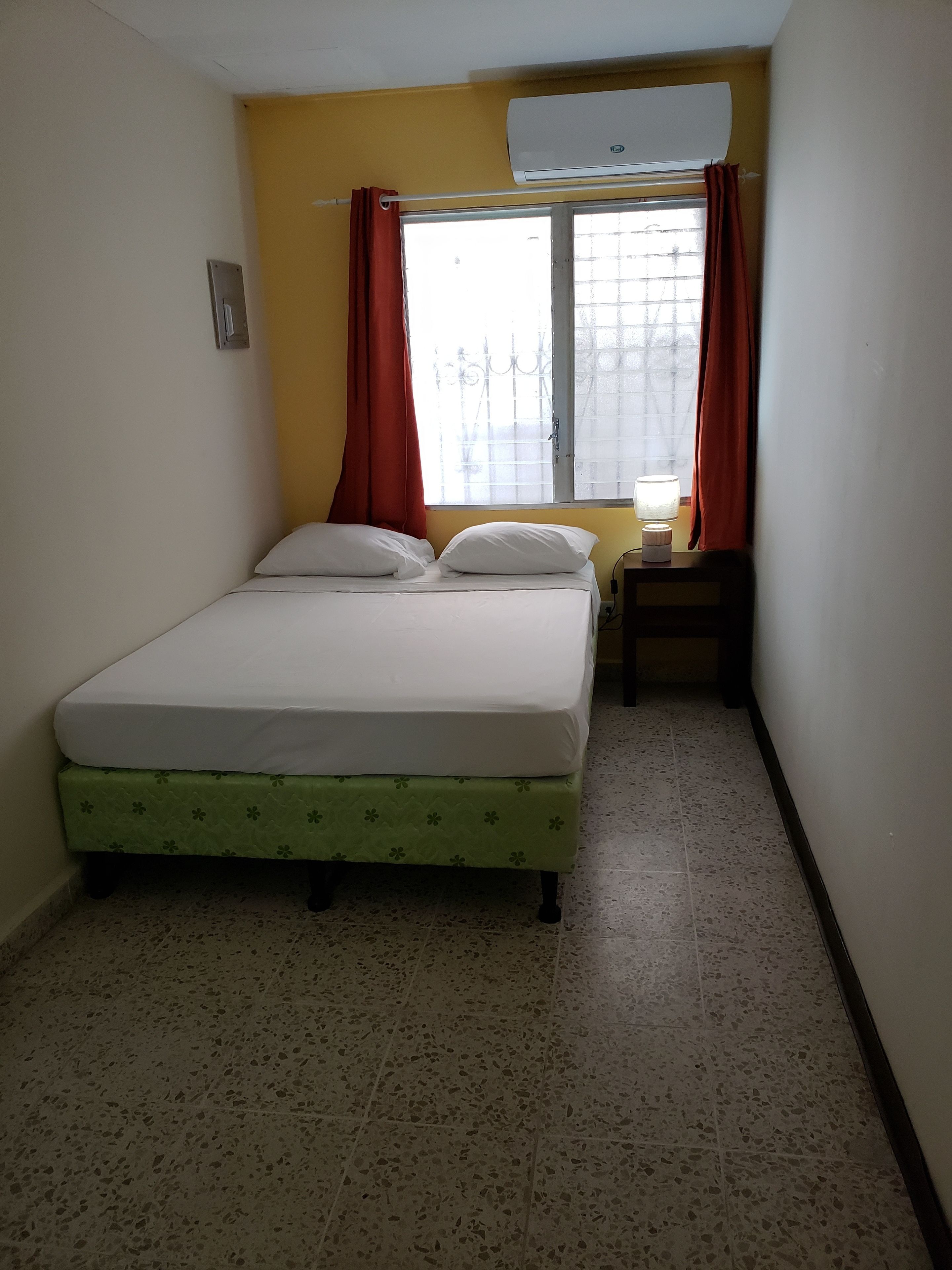 Una habitación pequeña con cama individual, cubrecama verde, ventana con cortinas rojas y un equipo de aire acondicionado encima.