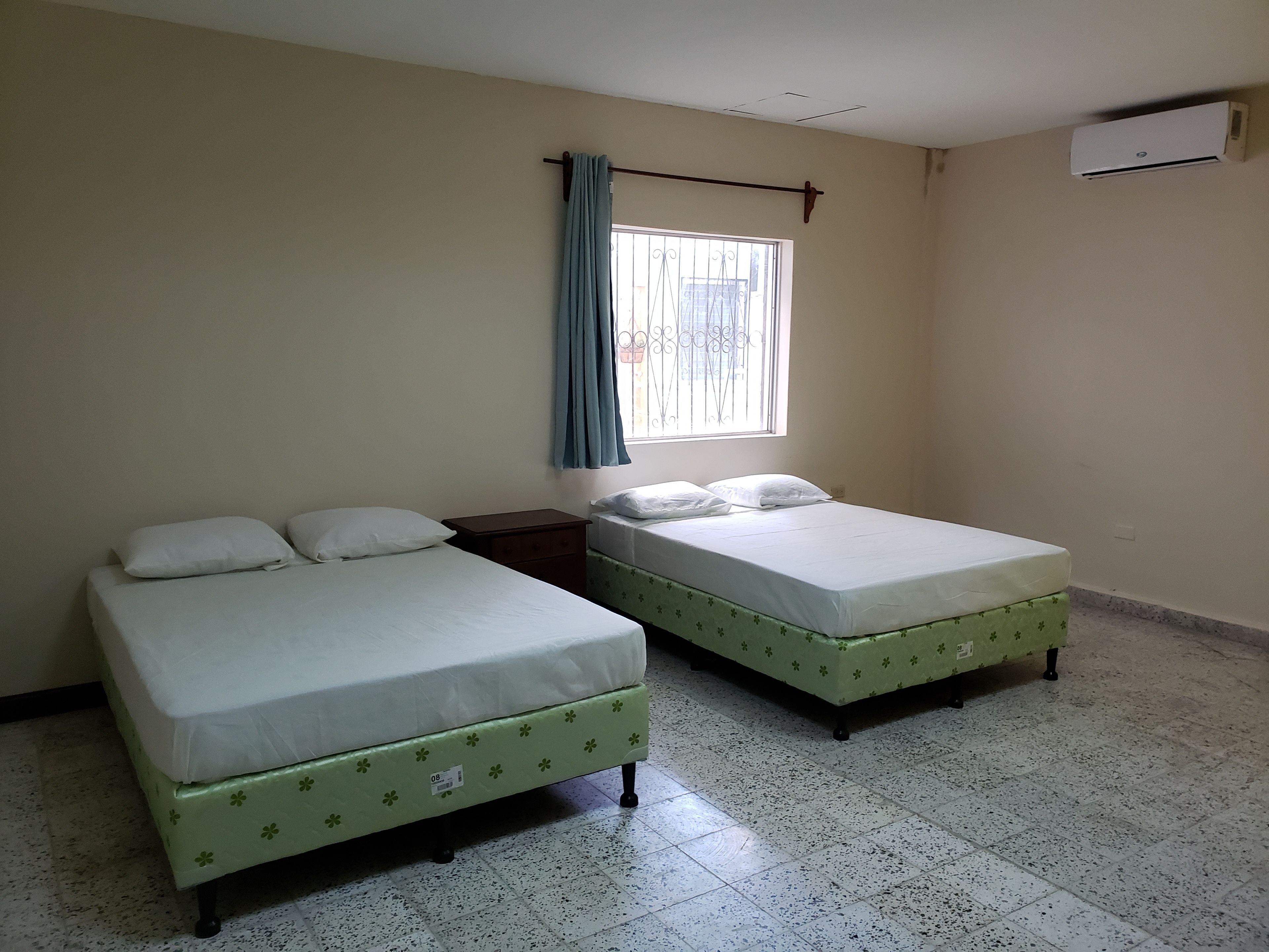 A room with two beds, a window with curtains, an air conditioning unit, and a tiled floor.