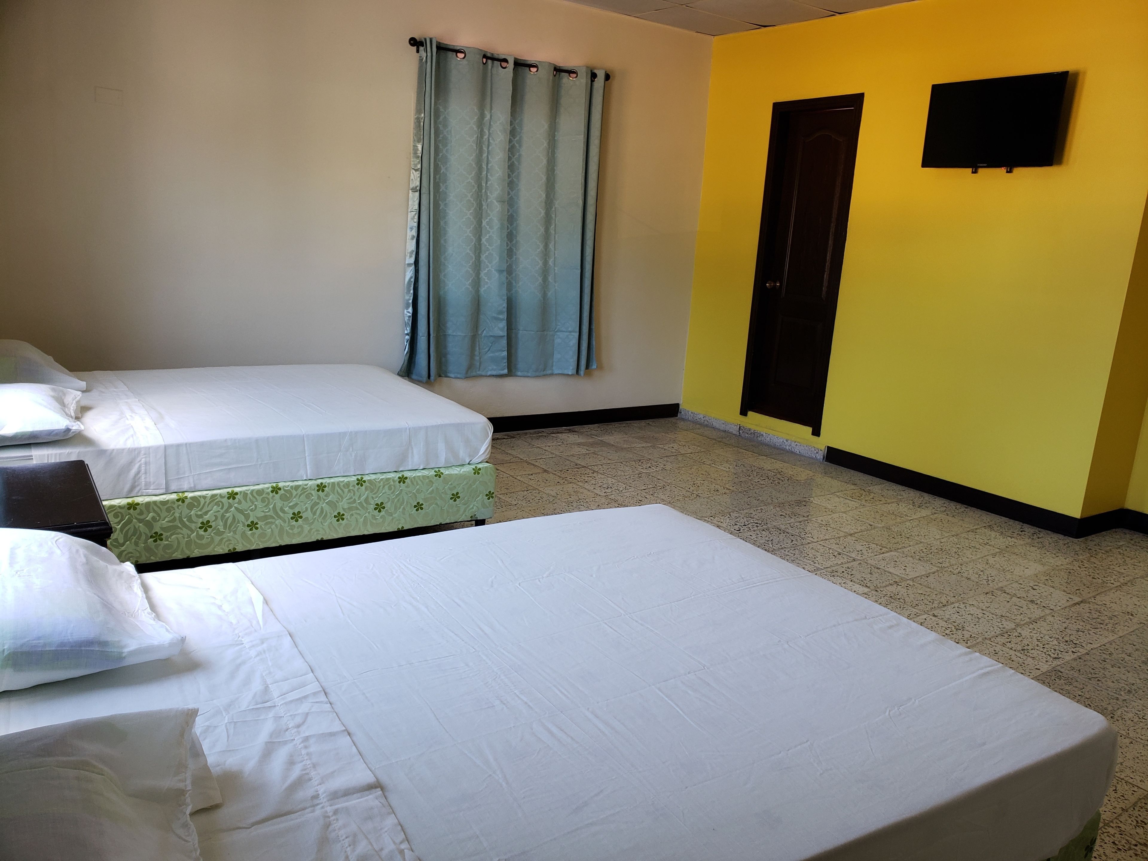 Quarto com duas camas, janela com cortinas, porta e televisão fixada na parede.
