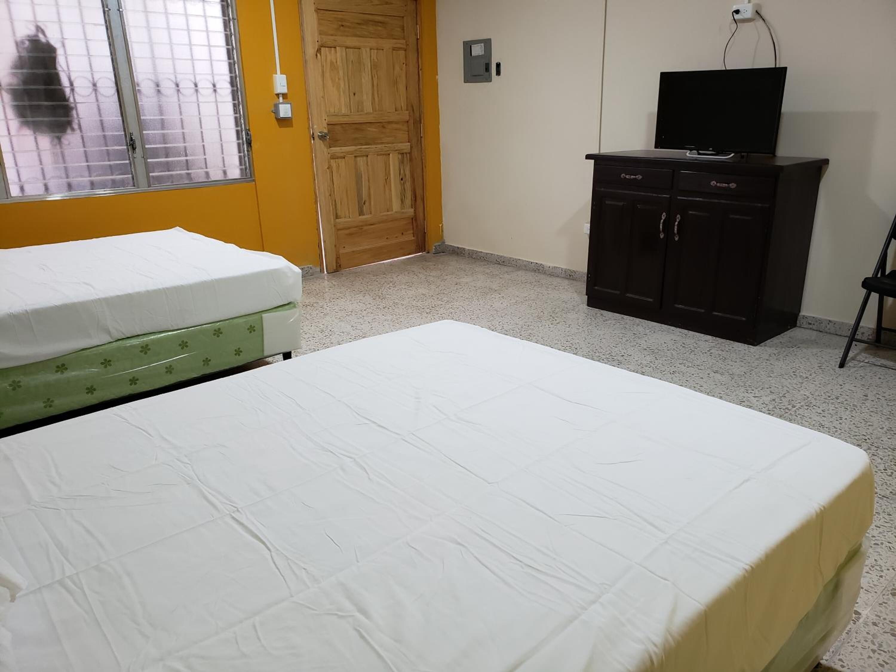 A room with a bed, a wooden door, a television on a cabinet, and a window with bars.