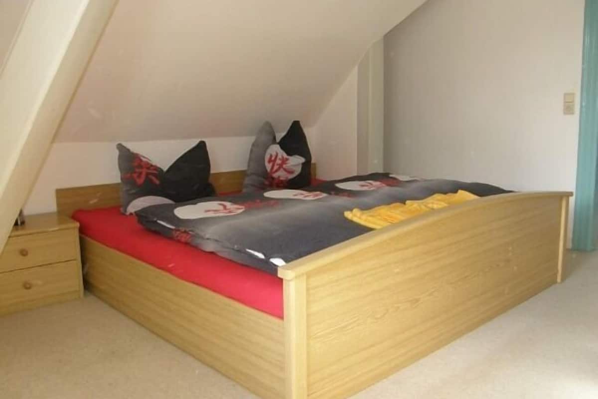 Quarto com teto inclinado, estrutura de cama de madeira, colchão vermelho e dois travesseiros com desenhos vermelhos e pretos.