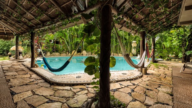 Un área de piscina con una hamaca, suelo de piedra y abundante vegetación.