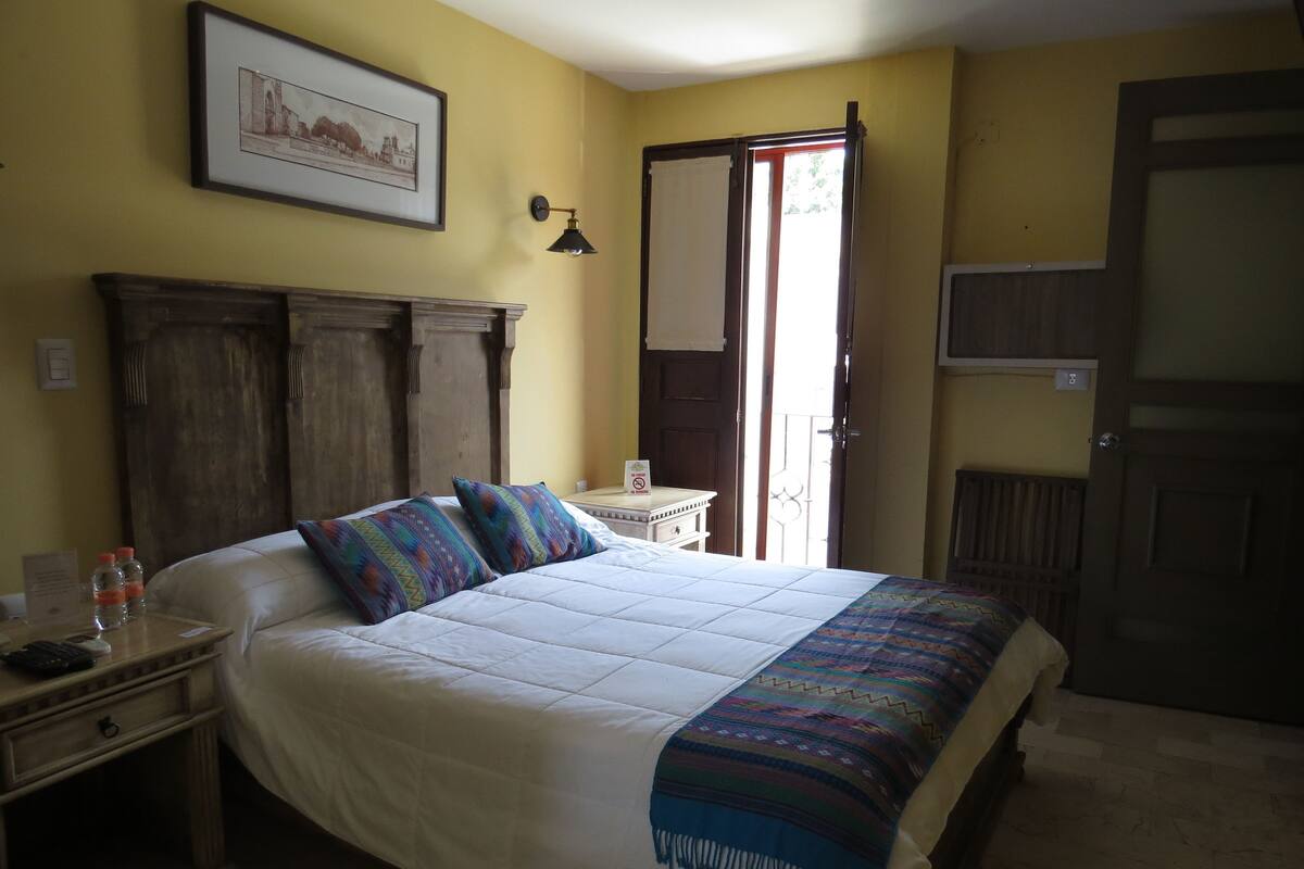Un dormitorio con una cama grande, una mesita de noche con lámpara, un cuadro enmarcado en la pared y una puerta que da a un balcón.