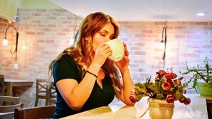 Eine Frau sitzt an einem Tisch mit einer weißen Tasse, einem Blumenstrauß in einer Vase und einer Ziegelwand im Hintergrund.