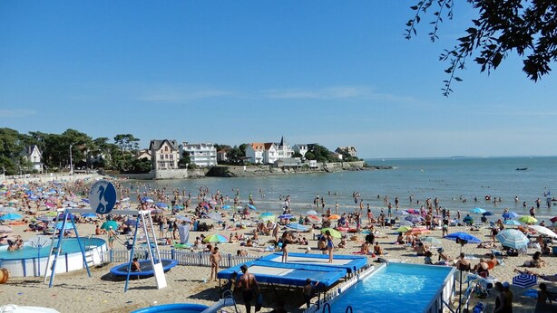 Une plage bondée, avec plusieurs piscines, des parasols et de nombreux vacanciers.