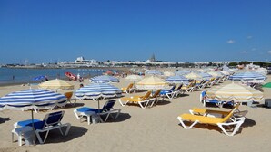Une plage bordée de rangées de chaises longues et de parasols, un ciel dégagé et une vue lointaine sur des bâtiments et une église.