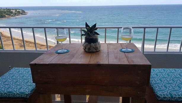 Ett träbord med två glas vitt vin och en ananas som mittpunkt på en balkong med utsikt över havet.