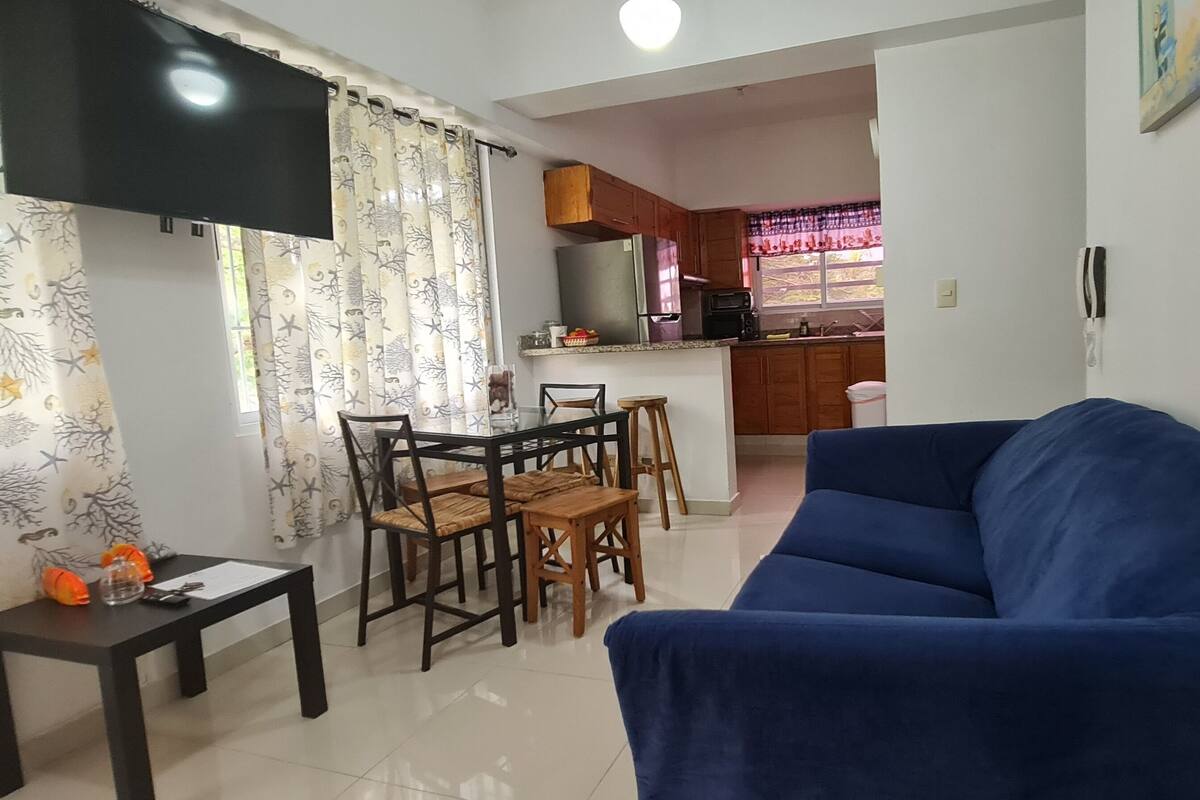 A living room with a blue sofa, a dining table with chairs, and a kitchen area in the background.