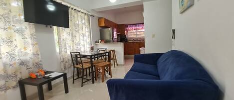 Una sala de estar con un sofá azul, una mesa de comedor con sillas y una cocina al fondo.