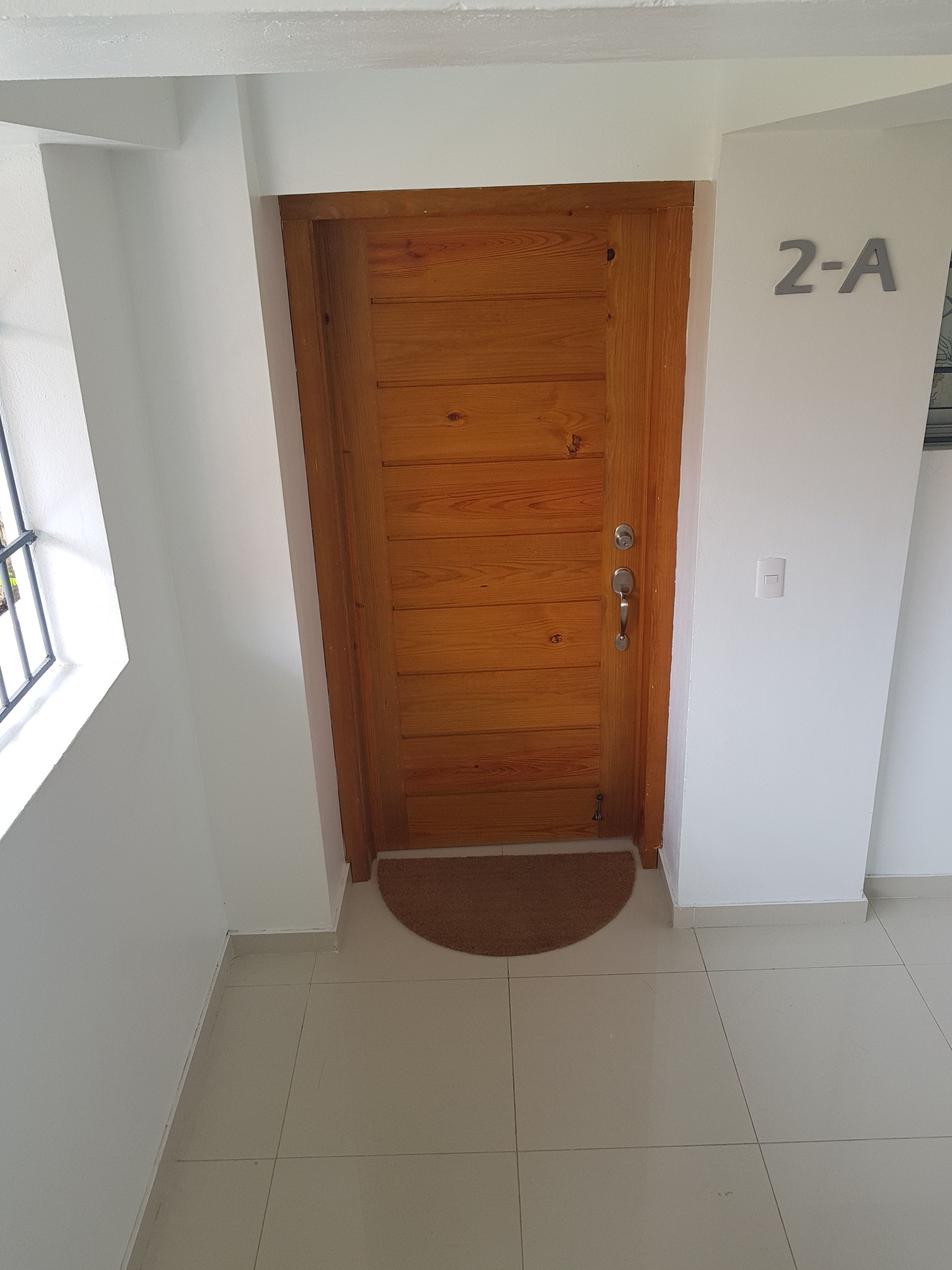 Una puerta de madera con manija de metal, un felpudo pequeño en la entrada y una pared con un letrero que dice "2-A".