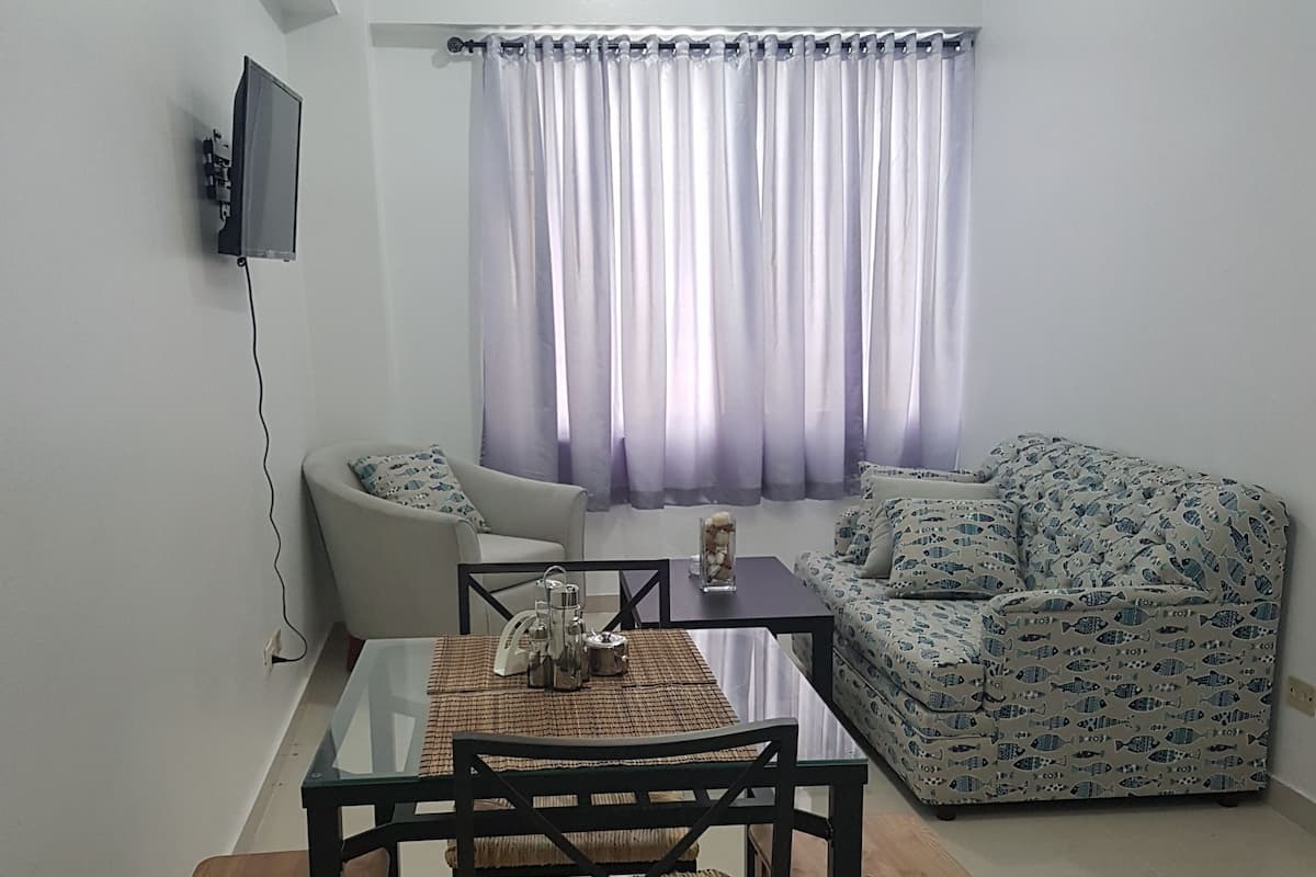 A living room with a glass dining table, two chairs, a sofa, and a TV mounted on the wall.